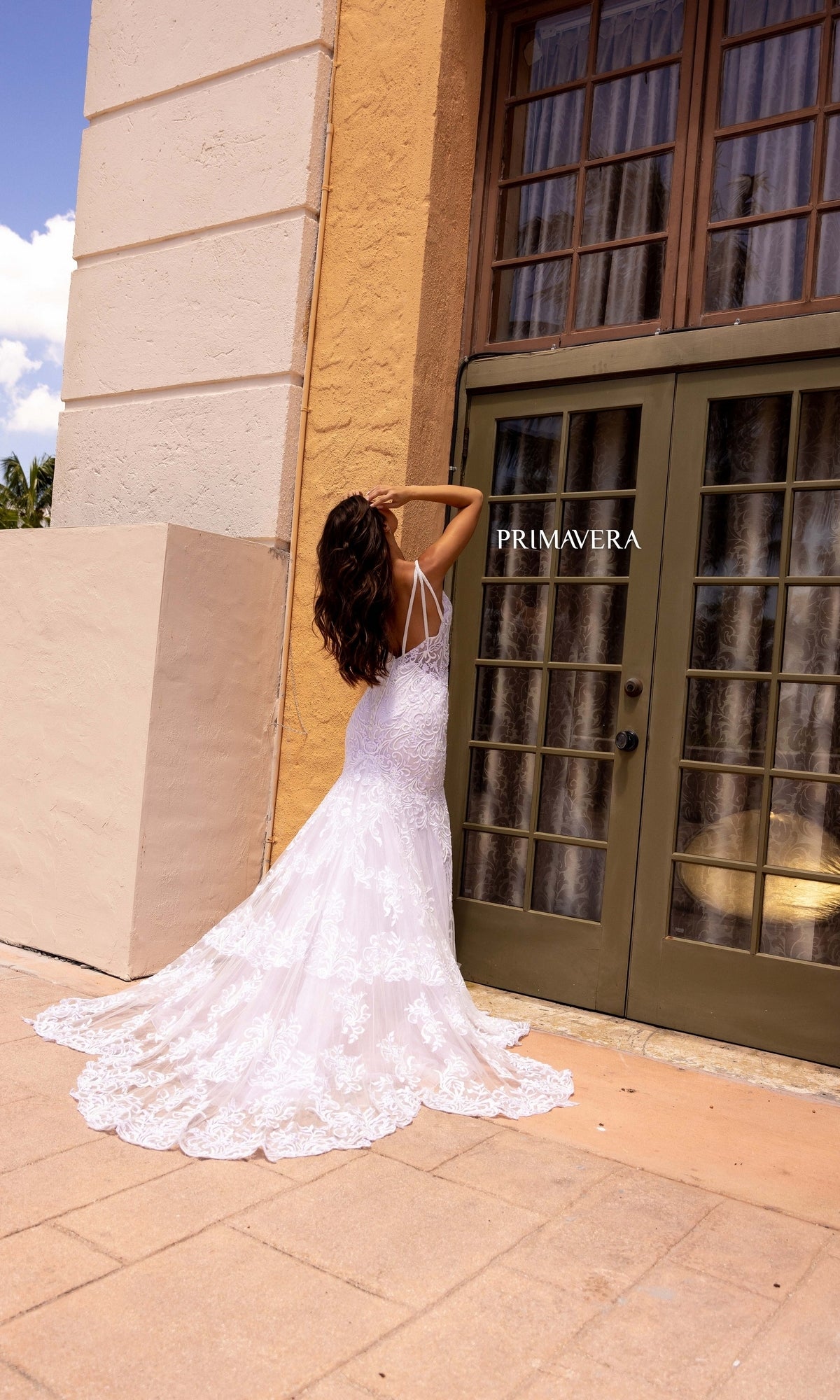 A woman stands outdoors with her back to the camera, wearing the Long Wedding Dress 11105 by Primavera—an ivory mermaid gown with lace embroidery—facing tall glass doors and a tan building.